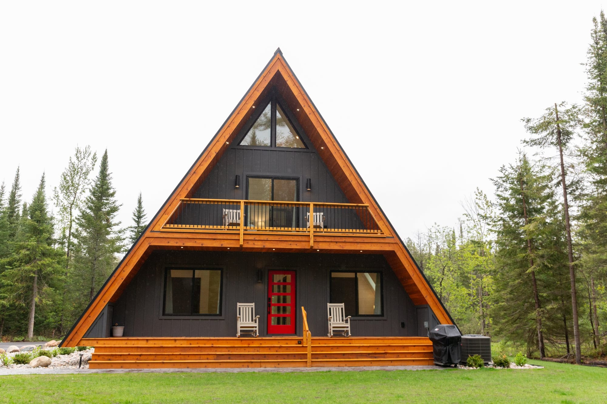 The A-frame from the front: charcoal siding, cedar trim, a red front door and a balcony under the gable